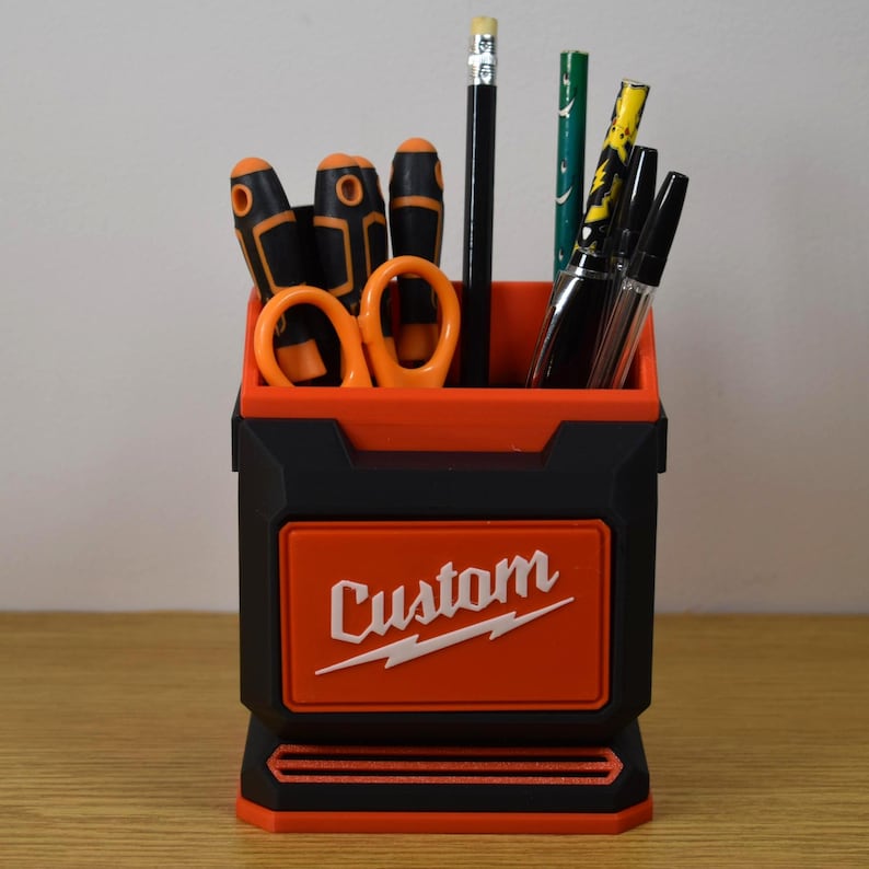 May include: Black and orange desk organizer with the word "Custom" in white. The organizer holds various tools, including screwdrivers, scissors, pencils, and pens. The design features a rectangular shape with a two-tone color scheme.