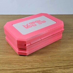 May include: A pink, rectangular box with a white lid. The lid has the word "Lego" embossed on it.