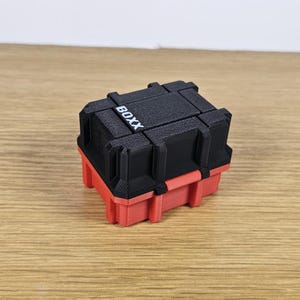 May include: A two-tone, red and black box with the word "BOXX" printed on the top. The box has a rectangular shape with a textured surface. The bottom half is red, and the top half is black.
