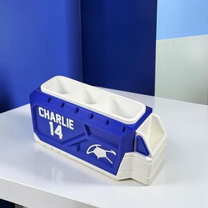 May include: A blue and white desk organizer featuring three rectangular compartments and one smaller side section. The side of the organizer is embossed with the name CHARLIE and the number 14, along with a stylized graphic icon of a sports jersey.