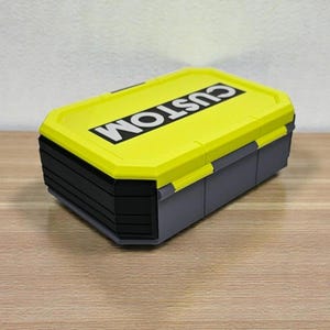 May include: A rectangular storage box with a bright yellow lid and gray base. The lid features the word "CUSTOM" in bold white letters. The box has a multi-tiered design and is suitable for organizing small items.