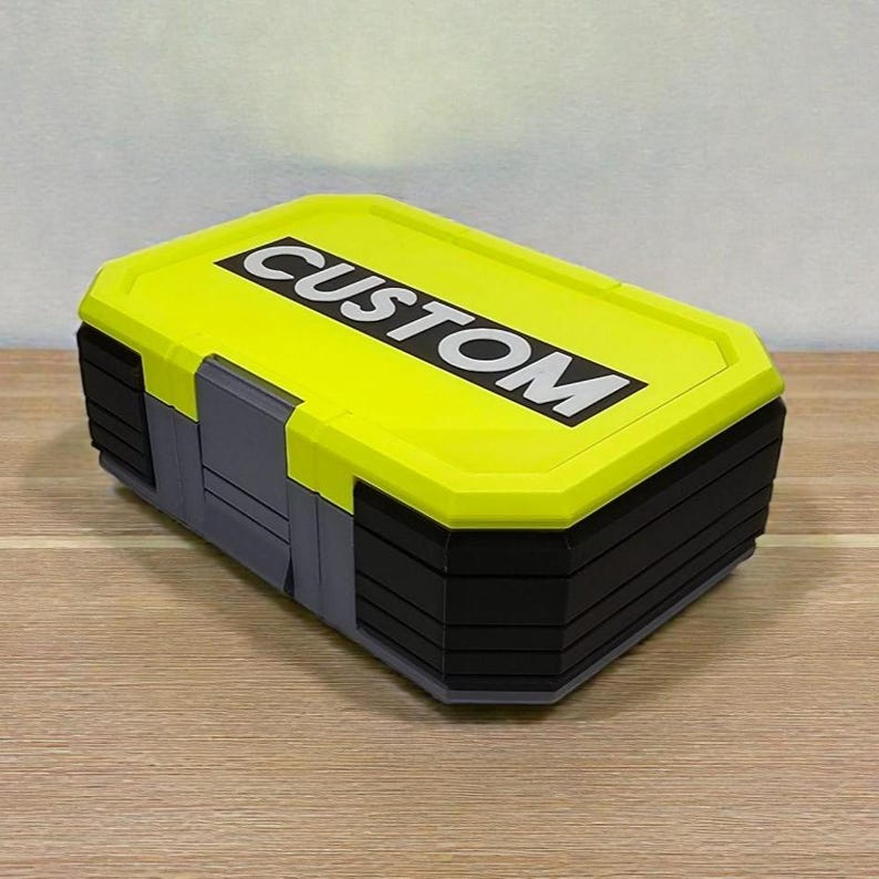 May include: A rectangular storage container with a bright yellow lid and black and gray body. The lid features the word "CUSTOM" in bold white letters. The container is sitting on a wooden surface.