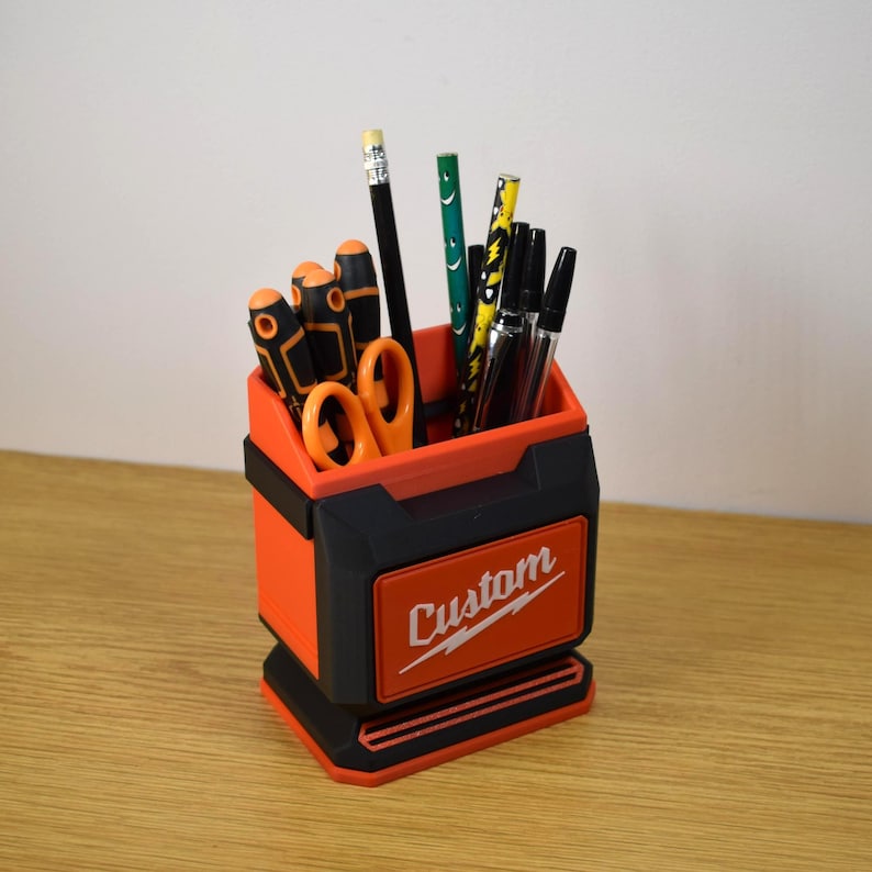 May include: An orange and black desk organizer with the word "Custom" on the front. The organizer holds various pens, pencils, and scissors. The design is reminiscent of a toolbox.