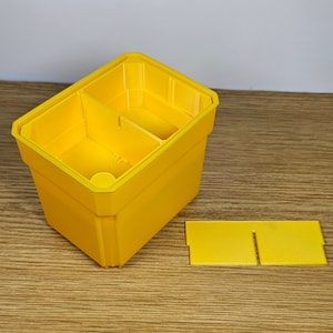 May include: A yellow plastic storage box with two compartments and a removable divider. The box is empty and sitting on a wooden surface.