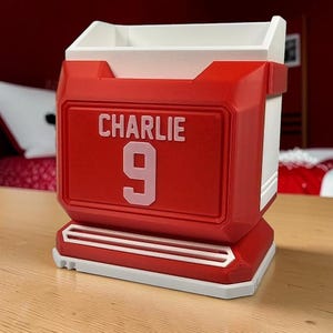 May include: A red and white desk organizer shaped like a sports jersey, featuring the name CHARLIE and the number 9 in light pink lettering on the front. The container has a rectangular open top and a structured base with horizontal white accents.