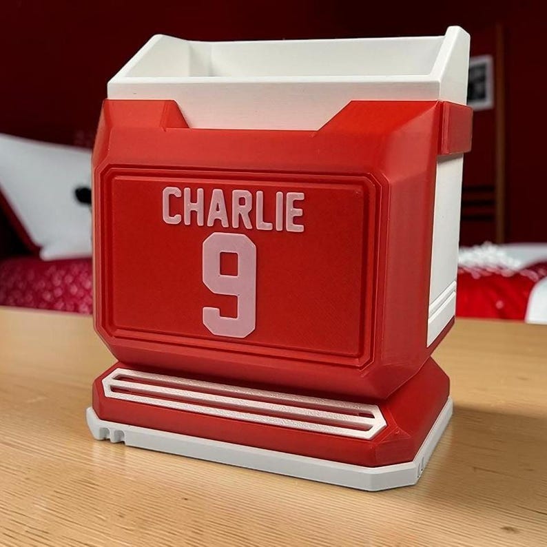May include: A red and white desk organizer shaped like a sports jersey, featuring the name CHARLIE and the number 9 in light pink lettering on the front. The container has a rectangular open top and a structured base with horizontal white accents.