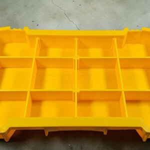 May include: Bright yellow storage organiser with multiple compartments. The organiser has a rectangular shape with a series of dividers creating individual storage spaces. Black accents are visible on the sides. The words "TOUGH SYSTEM" are printed on the side.