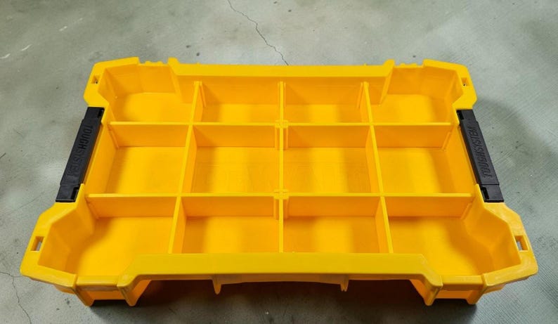 May include: Bright yellow storage organiser with multiple compartments. The organiser has a rectangular shape with a series of dividers creating individual storage spaces. Black accents are visible on the sides. The words "TOUGH SYSTEM" are printed on the side.