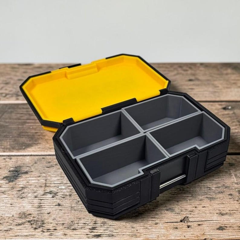 May include: A black and yellow organizer box with a hinged lid. The open box reveals four gray compartments. The lid is yellow, and the box has a black exterior with gray trim. The box is on a wooden surface.