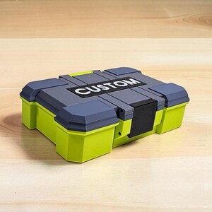 Peut inclure: Une bo&icirc;te de rangement rectangulaire avec un couvercle gris et des c&ocirc;t&eacute;s vert citron. Le mot "CUSTOM" est imprim&eacute; en blanc sur le dessus. La bo&icirc;te est pos&eacute;e sur une surface en bois brun clair.