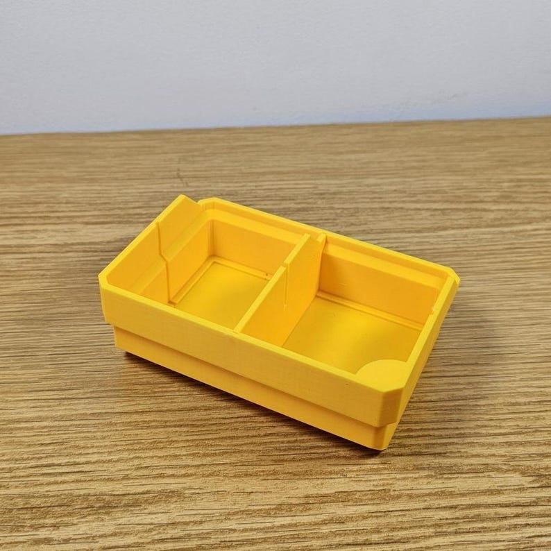 May include: A bright yellow, rectangular storage container with two internal dividers. The container is made of a solid material and has a smooth finish. The design is simple and functional, suitable for organizing small items.