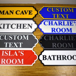 Puede incluir: Un conjunto de letreros decorativos con textos como "Man Cave", "Kitchen", "Custom Text", "Isla's Room", "Charlie's Room" y "Bathroom". Los letreros presentan diferentes combinaciones de colores, como amarillo, blanco, negro, rojo y azul.