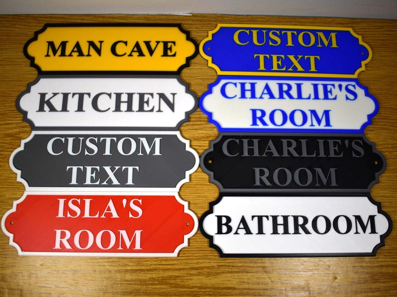 Puede incluir: Un conjunto de letreros decorativos con textos como "Man Cave", "Kitchen", "Custom Text", "Isla's Room", "Charlie's Room" y "Bathroom". Los letreros presentan diferentes combinaciones de colores, como amarillo, blanco, negro, rojo y azul.