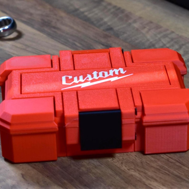 May include: A red plastic tool case with the word "Custom" in white script. The case has a black latch and two side compartments. The case is sitting on a wooden surface.
