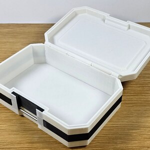 Puede incluir: Una caja de pl&aacute;stico blanca con un borde negro. La caja est&aacute; abierta y el interior est&aacute; vac&iacute;o.