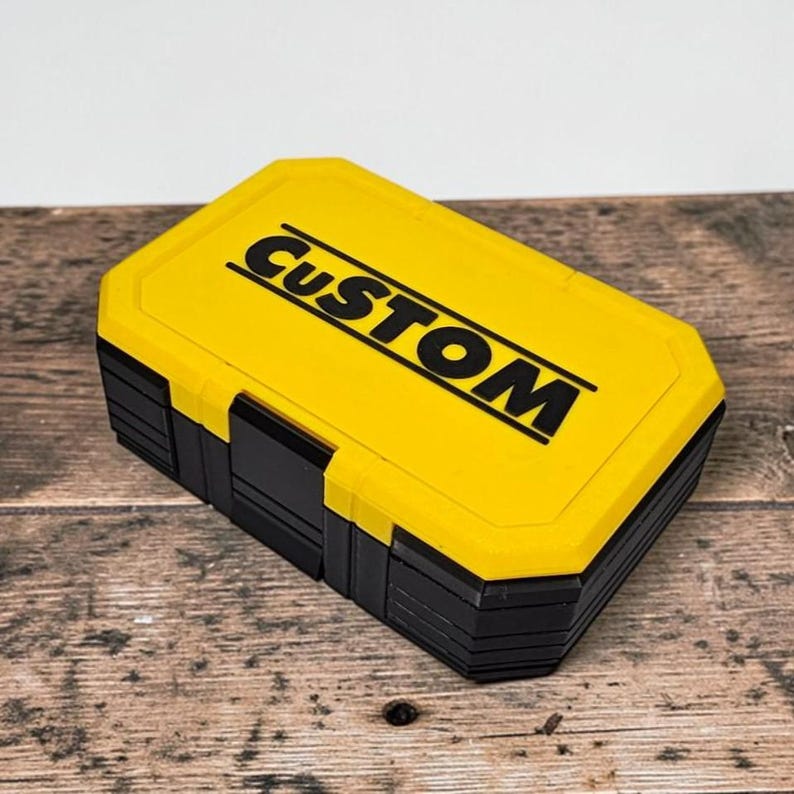 May include: A yellow and black rectangular box with the word "CUSTOM" printed in black on the lid. The box is sitting on a wooden surface.