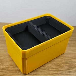 May include: A yellow plastic storage box with two compartments. The compartments are lined with black plastic.