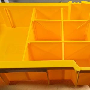 May include: A bright yellow plastic storage organizer tray with multiple fixed compartments of varying sizes. The unit features a black latch on the right side with the text TOUGHSYSTEM embossed on it, designed for modular tool storage organization.