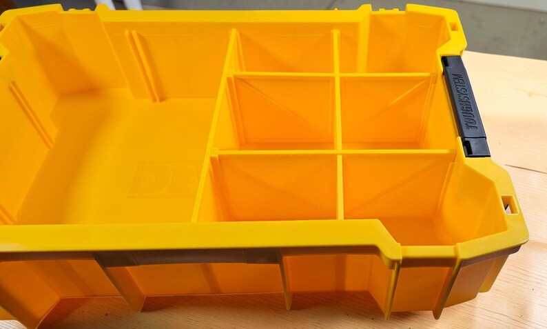 May include: A bright yellow plastic storage organizer tray with multiple fixed compartments of varying sizes. The unit features a black latch on the right side with the text TOUGHSYSTEM embossed on it, designed for modular tool storage organization.