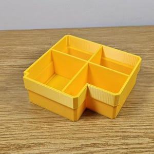 May include: A yellow plastic organizer with six compartments. The organizer has a stepped design and is made of a solid, bright yellow material. The compartments are divided by vertical walls.