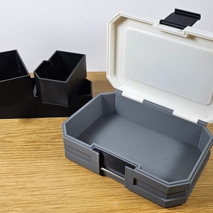 K&ouml;nnte beinhalten: Eine wei&szlig;e und graue Plastik-Aufbewahrungsbox mit einem Scharnierdeckel. Die Box ist ge&ouml;ffnet und das Innere ist grau. Auf der linken Seite des Bildes befinden sich vier kleinere schwarze Plastikboxen, die &uuml;bereinander gestapelt sind.