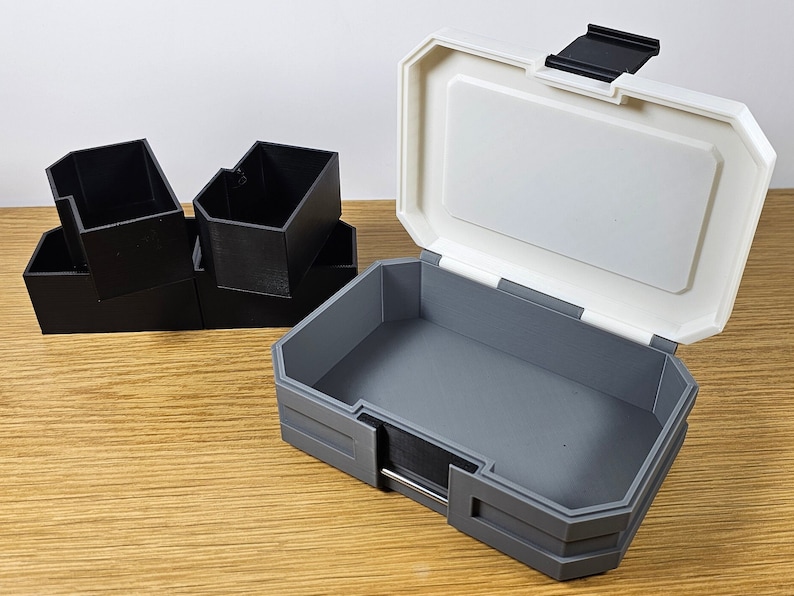 K&ouml;nnte beinhalten: Eine wei&szlig;e und graue Plastik-Aufbewahrungsbox mit einem Scharnierdeckel. Die Box ist ge&ouml;ffnet und das Innere ist grau. Auf der linken Seite des Bildes befinden sich vier kleinere schwarze Plastikboxen, die &uuml;bereinander gestapelt sind.