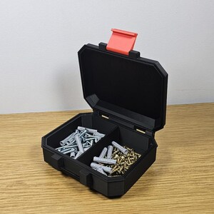 May include: A black, open toolbox with a red latch, filled with screws and wall anchors. The toolbox is divided into three compartments, each containing different types of hardware. The toolbox is sitting on a wooden surface.