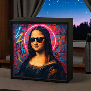 Peut inclure: Une boîte à lumière encadrée de noir avec une image colorée de la Joconde portant des lunettes de soleil. L'arrière-plan est un mélange vibrant de rose, de bleu et de jaune.