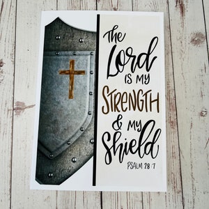May include: A white card with a silver shield and a cross on it. The text on the card reads "The Lord is my strength & my shield Psalm 28:7".