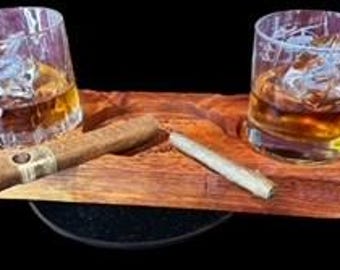Puede incluir: Dos vasos de whisky con hielo y líquido ámbar sobre una bandeja de madera, junto a un cigarro y un cigarrillo. La bandeja tiene dos hendiduras circulares para los vasos y una ranura para el cigarro y el cigarrillo. El fondo es negro.