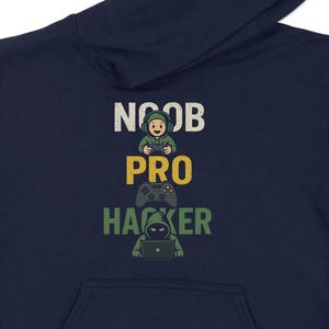 May include: Navy blue hoodie with a front pocket and hood. The front features the words "NOOB", "PRO", and "HACKER" stacked vertically, each with a corresponding cartoon graphic of a gamer. The hoodie is made of a soft material.