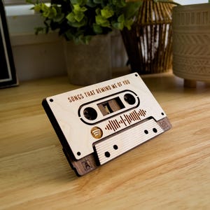 Puede incluir: Diseño de cinta de cassette de madera con el texto "SONGS THAT REMIND ME OF YOU". El diseño incluye un logotipo de Spotify y un gráfico de onda de sonido. La cinta de cassette está hecha de madera clara.