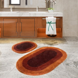 May include: Two brown and orange oval bath mats with a darker brown center. The mats are in a bathroom with a white toilet and a wooden cabinet.