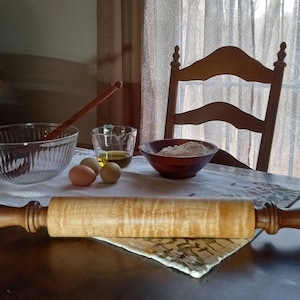 May include: A wooden rolling pin with a light brown maple wood body and dark brown wooden handles. The rolling pin is on a table with a white tablecloth, next to a bowl of flour, a bowl of olive oil, and three eggs.