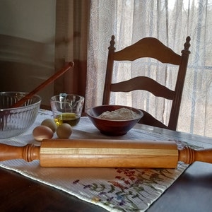 May include: A wooden rolling pin with a light wood center stripe, resting on a white tablecloth with a floral pattern. There are three eggs, a bowl of flour, a glass bowl with olive oil, and a wooden spoon in the background.