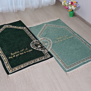 Puede incluir: Dos alfombras de oración, una verde oscuro y otra turquesa, con bordes florales y geométricos. Cada alfombra tiene un diseño central y texto dorado. La alfombra verde oscuro dice "Ibrahim" y la turquesa dice "Sara".