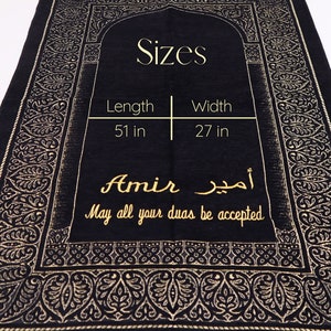 May include: Black prayer rug with gold embroidery featuring a traditional Islamic design. The rug measures 51 inches long and 27 inches wide. It has the name "Amir" and the Arabic phrase "أمير" written in gold. The text "May all your duas be accepted" is also written in gold.