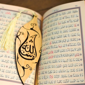 May include: A wooden bookmark shaped like a leaf with intricate calligraphy in black ink. The bookmark features the Arabic word "Allah" and has a cream-colored tassel.