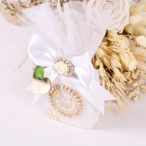 May include: White favor bag with a white tulle overlay and a white satin bow. The bag has a gold design with rhinestones and a heart-shaped tag that says "Mawwalid  2023".