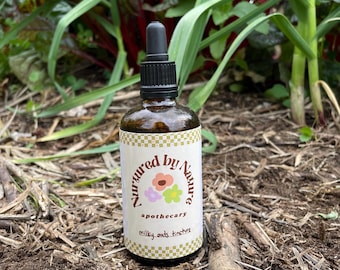 Könnte beinhalten: Eine braune Apothekerflasche aus Glas mit schwarzer Pipette, beschriftet mit "Nurtured by Nature". Das Etikett zeigt Blumenillustrationen und den Text "milky oats tincture". Die Flasche steht auf einer Mulchschicht mit grünen Pflanzen im Hintergrund.