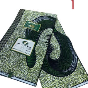 Può includere: Un tessuto piegato con un motivo verde e giallo, con un grande disegno a spirale verde scuro. Il tessuto ha un motivo ripetuto di piccole forme gialle. Il testo "PRINTED BY VUSCO IN HOLLAND" è visibile su un'etichetta.