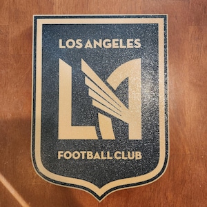 May include: A shield-shaped emblem with the text "LOS ANGELES" at the top and "FOOTBALL CLUB" at the bottom. The center features a stylized "LA" logo in gold against a black background. The emblem has a textured appearance.
