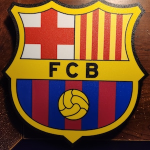 May include: A yellow and black shield-shaped logo with the text "FCB" in black letters. The shield has a red and yellow striped section, a white section with a red cross, and a blue and red striped section with a yellow soccer ball.