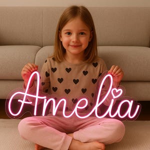 May include: A pink neon sign that spells out the name "Amelia" in a cursive font, with a heart-shaped detail. The sign is held by a young girl wearing a heart-patterned shirt and pink pants.