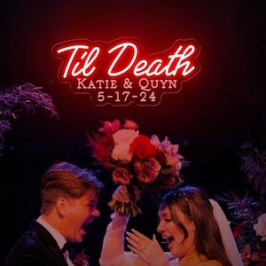 Op de afbeelding: Een huwelijksceremonie met een stel dat onder een neonbord staat met de tekst "Til Death Katie & Quyn 5-17-24". De bruid draagt een witte jurk en sluier, en de bruidegom draagt een zwart pak. De bruid houdt een boeket rode en roze bloemen vast.
