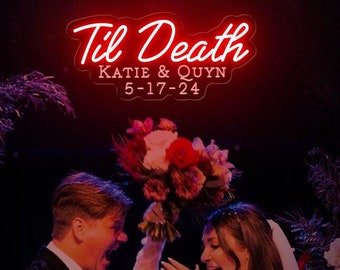 May include: A wedding ceremony with a couple standing under a neon sign that reads "Til Death Katie & Quyn 5-17-24". The bride is wearing a white dress and veil, and the groom is wearing a black suit. The bride is holding a bouquet of red and pink flowers.