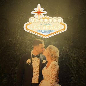 K&ouml;nnte beinhalten: Ein k&uuml;ssendes Paar vor einem Schild mit der Aufschrift "Married in Fabulous Las Vegas". Das Schild ist im Stil des ber&uuml;hmten Las Vegas-Schildes gehalten, mit einem Stern oben. Es zeigt auch die Namen "Mr. & Mrs. Brown" und das Datum "May 13, 2025."