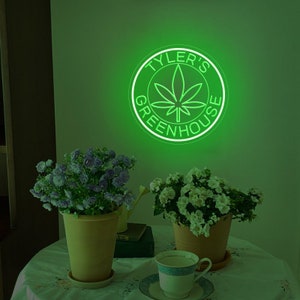 Puede incluir: Un letrero de ne&oacute;n verde con un c&iacute;rculo alrededor que dice "Tyler's Greenhouse" con una hoja de marihuana dentro del c&iacute;rculo. El letrero est&aacute; en una pared verde con dos plantas en macetas delante.
