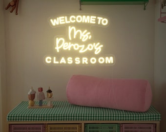 May include: White neon sign with the text "Welcome to Ms. Perozo's Classroom". The sign is hanging on a wall above a pink pillow and a wooden shelf with colorful toys.