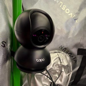 Puede incluir: Una cámara de seguridad Tapo negra con una parte superior e inferior redondeadas. La cámara tiene una lente y una pequeña luz roja. El logotipo de Tapo es visible en la base. La cámara está montada en un poste verde.